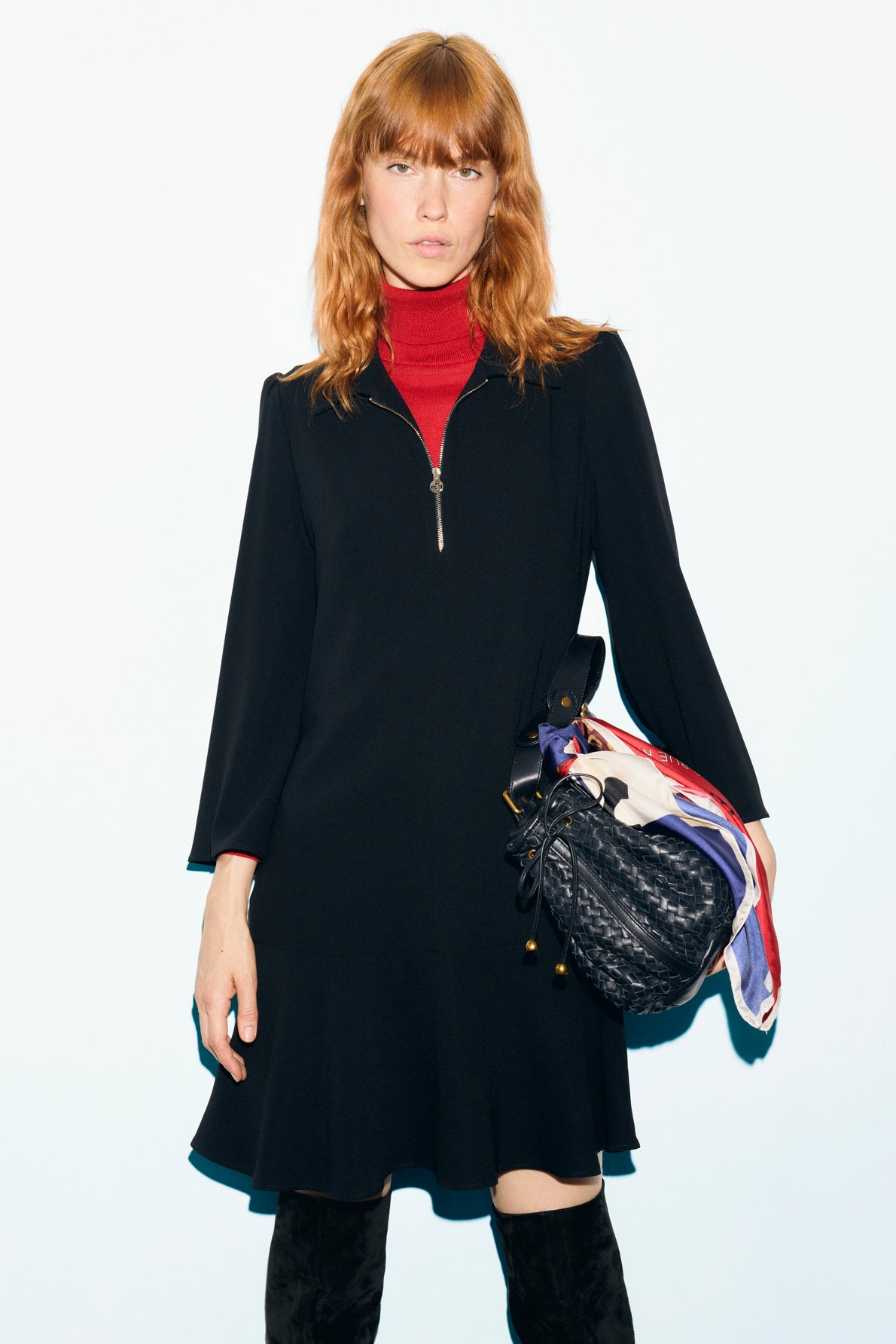 A woman with reddish hair in a crêpe noir robe courte and red turtleneck holds a black handbag with colorful scarves tied to it, standing against a plain light background.