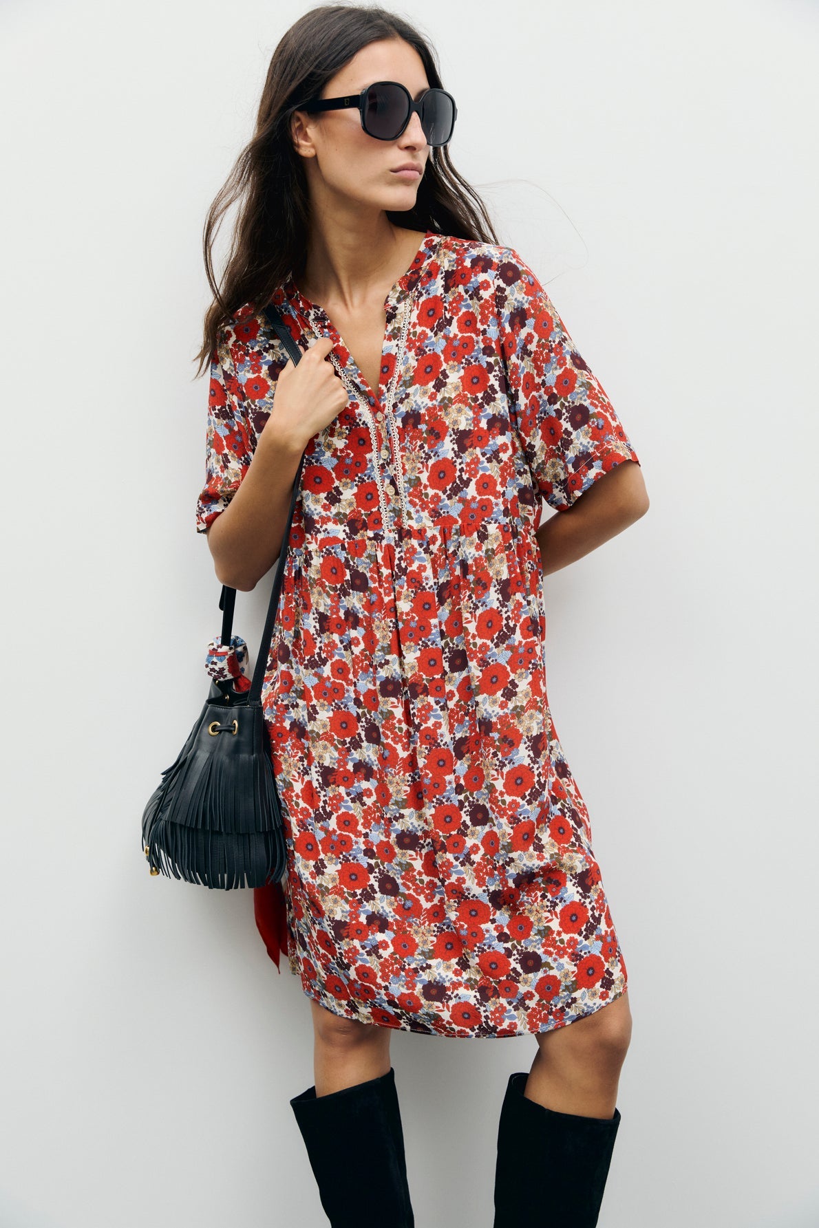 Woman wearing a loose, robe fleurie courte with red and white flowers in an empire waist (taille empire) cut, large black sunglasses, a black fringe handbag, and knee-high black boots, standing against a plain white background.