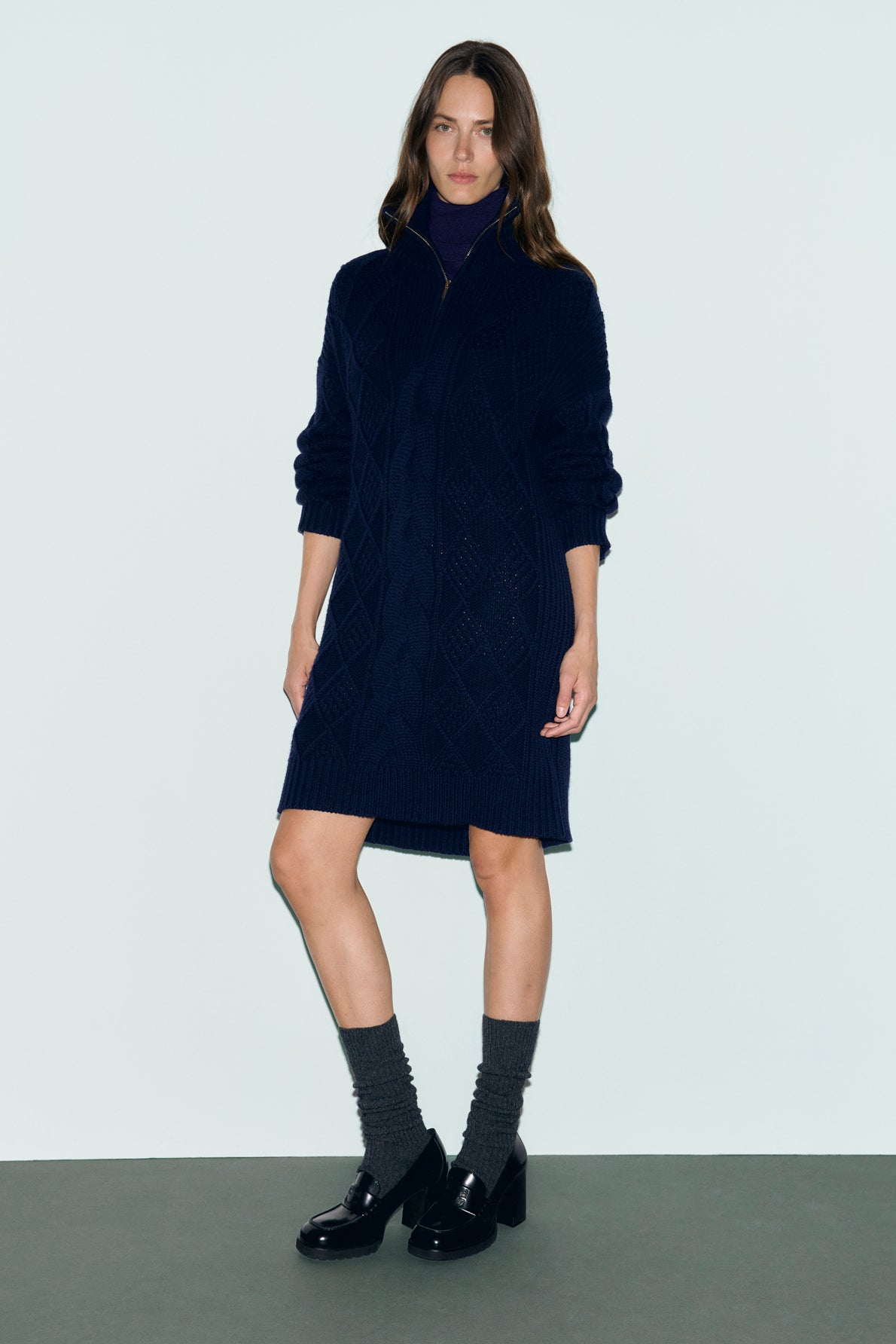 A woman stands against a plain background wearing a navy blue robe pull laine, paired with gray socks and black heeled loafers. She has long brown hair and a neutral expression, with her arms relaxed by her sides.