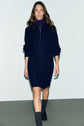 A woman wearing a navy blue robe pull laine courte with a col camionneur and front zipper, black shoes, and gray socks walks on a gray floor against a light background.