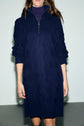 A person wears a navy blue, cable-knit robe pull laine with a col camionneur zip-up collar and long sleeves. The robe courte reaches above the knees. The person’s head is partially out of frame.