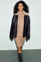 A woman with curly hair wears a robe col roulé in a tan turtleneck sweater dress, paired with a dark brown leather jacket featuring a shearling collar and stylish brown boots, standing against a light background.