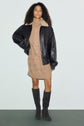 A woman wearing a brown leather jacket with a shearling collar, a beige robe pull laine torsadée, and knee-high suede boots stands against a plain light background.
