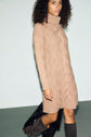 Woman with curly hair wearing a tan robe torsadée col roulé sweater dress and dark knee-high boots stands against a light background, holding a black coat at her side.