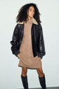 A person with curly hair wears a brown robe pull laine, paired with a black shearling-collar leather jacket and knee-high dark boots, standing against a plain light background.
