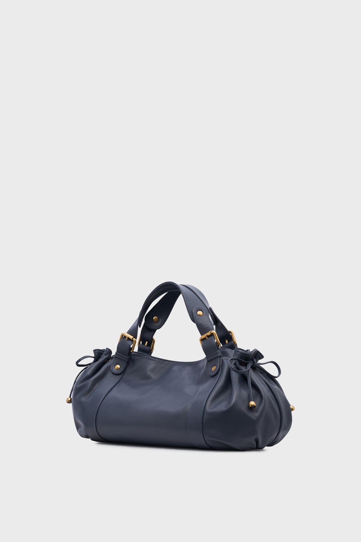 A dark blue leather sac à main cuir with gold hardware, two short handles, and gathered sides adorned with small bows, set against a plain light gray background—a timeless piece from la maroquinerie Darel reminiscent of the classic sac 24H.