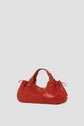 A red leather sac à main with gold hardware, double handles, and side drawstring details is displayed against a plain light gray background, reflecting the timeless elegance of maroquinerie Darel.