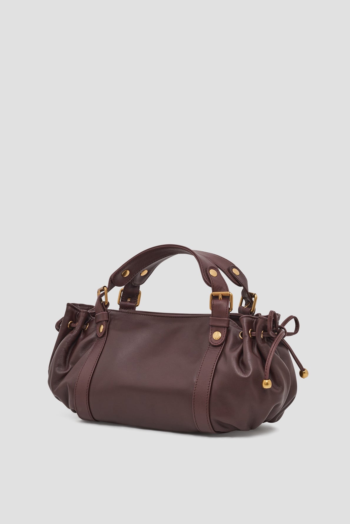 A dark brown cuir sac à main in a format mini, featuring gold hardware, two handles, side drawstring details, and a slightly gathered design, set against a plain light gray background.