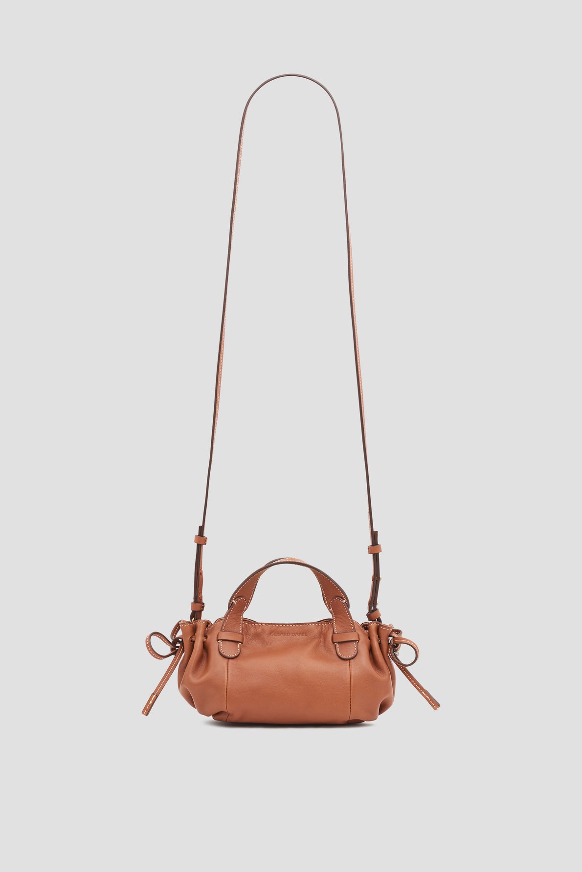 A small brown sac bandoulière cuir, part of the exclusive collaboration Merci Micro 24 collection, with short handles, an adjustable long crossbody strap, and side ties, displayed against a light gray background.
