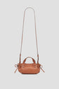 A small brown sac bandoulière cuir, part of the exclusive collaboration Merci Micro 24 collection, with short handles, an adjustable long crossbody strap, and side ties, displayed against a light gray background.