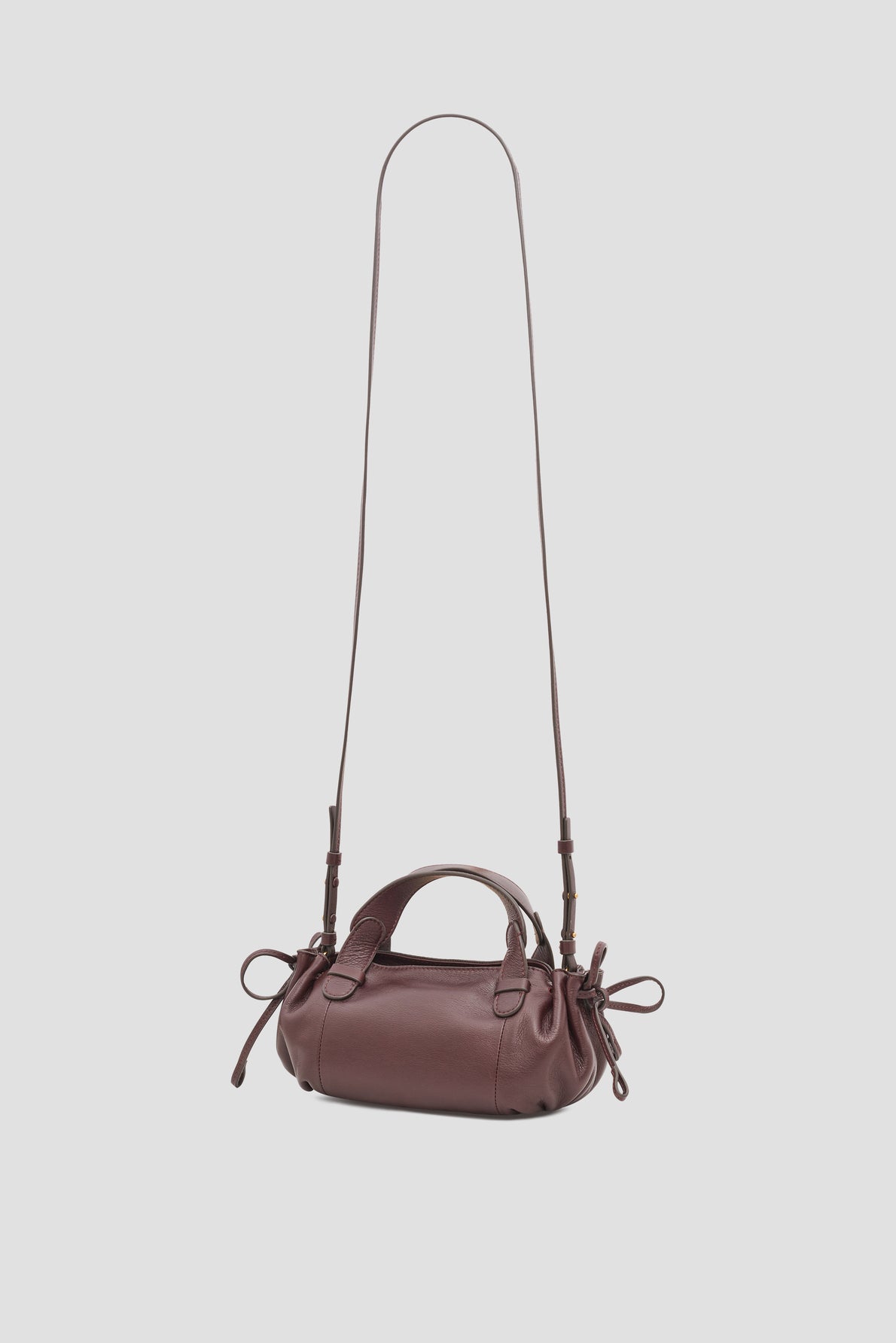 A small, dark brown cuir leather sac bandoulière handbag with a long shoulder strap, two short handles, and side drawstring details, displayed against a plain light gray background.