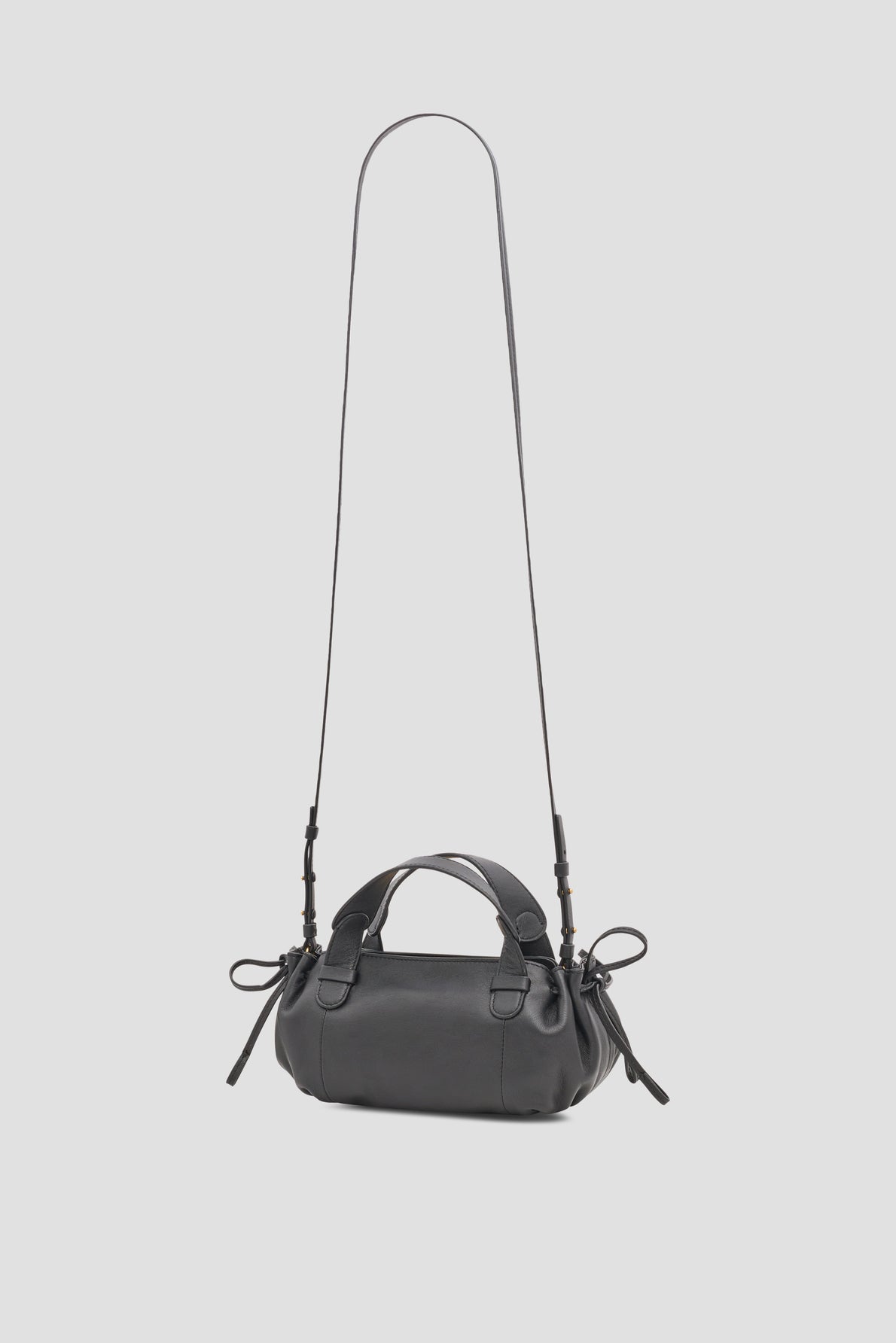 A small black sac bandoulière cuir, crafted in collaboration Merci, with short handles and a long adjustable shoulder strap, featuring decorative knots on each side, displayed against a light gray background.