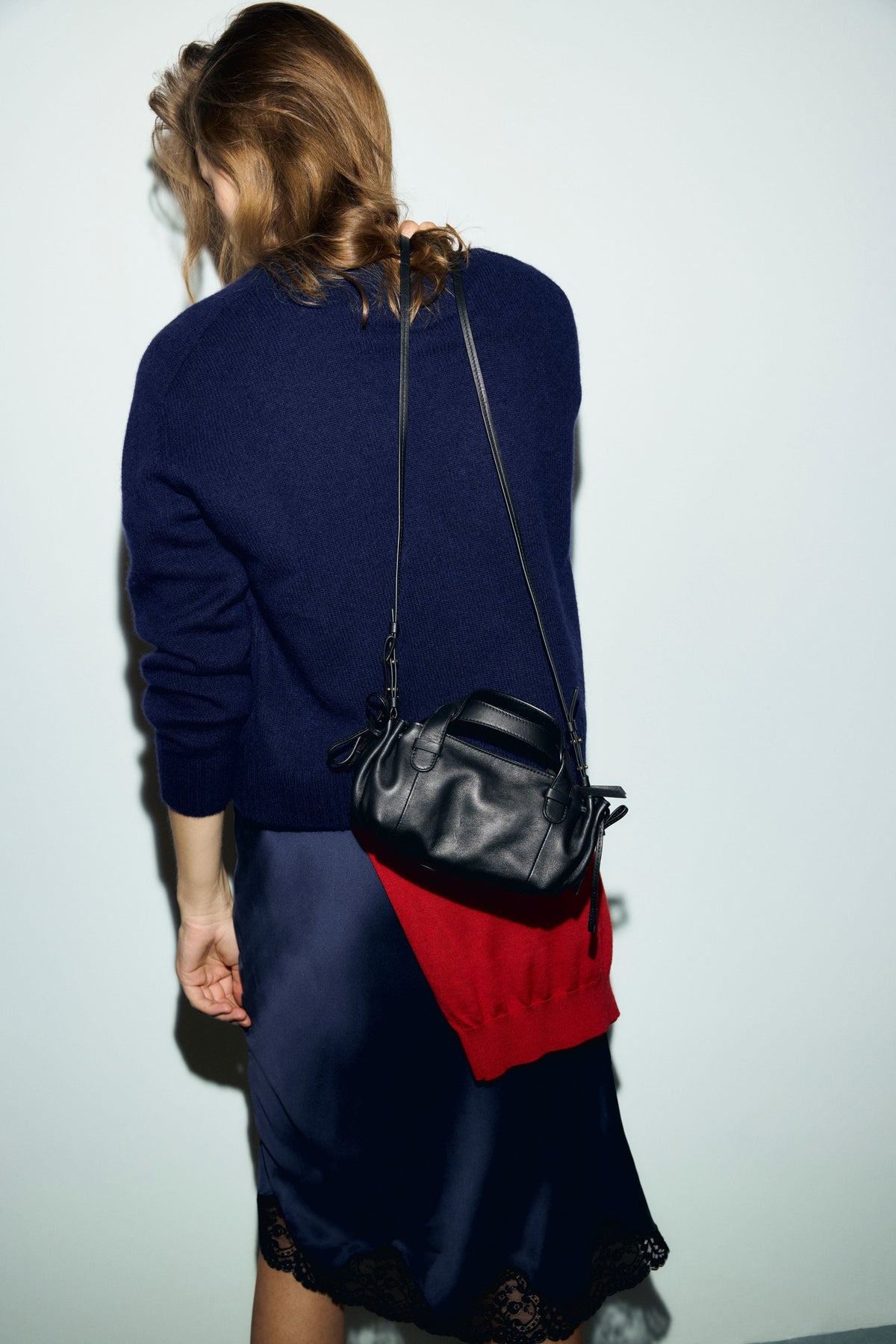 A woman with wavy hair faces away, wearing a navy sweater and a satin skirt with lace trim. She carries a sac bandoulière cuir Micro 24, along with a red garment draped over her arm—a chic look inspired by the collaboration Merci.