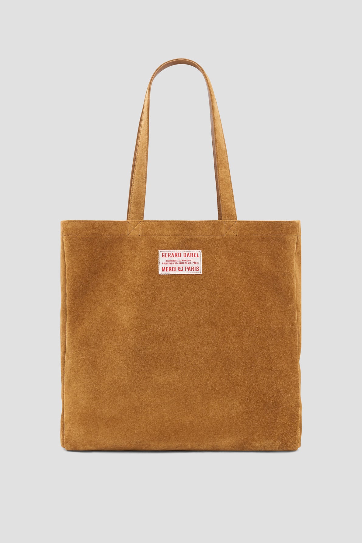 Brown suede tote bag cuir with two straps and a rectangular patch near the top center that reads Gérard Darel Merci Paris in red text on a white background. The background is plain and light gray.