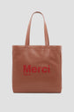 A brown leather tote bag cuir with two handles and the words Merci Paris printed in red on the front. The design captures the effortless elegance of Gérard Darel, set against a plain, light gray background.