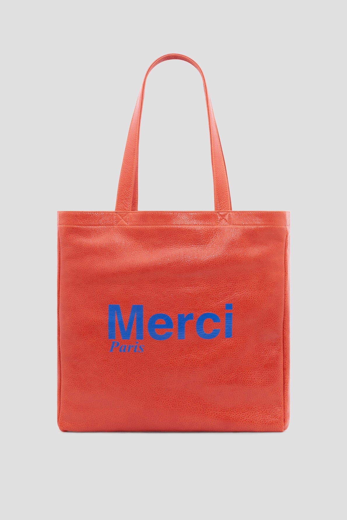 A bright red tote bag with two handles, inspired by Gérard Darel’s chic style. Merci Paris is printed on the front in large blue letters. The background is plain light gray.