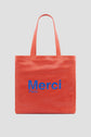 A bright red tote bag with two handles, inspired by Gérard Darel’s chic style. Merci Paris is printed on the front in large blue letters. The background is plain light gray.
