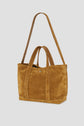 Brown suede tote bag, crafted in cuir velours, with two short handles and one long shoulder strap, featuring visible white stitching and a small metallic logo detail on the front. The background is plain light gray.