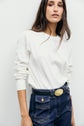 A woman with long brown hair wears a white tee-shirt Bara à manches longues en coton épais and dark blue jeans with a black belt, standing against a plain white background.