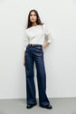 A woman stands against a plain white wall, wearing a white tee-shirt Bara à manches longues en coton épais, high-waisted dark blue wide-leg jeans, a black belt, and black shoes. She has long brown hair and a neutral expression.