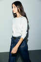 A woman with long dark hair, wearing a white tee-shirt Bara à manches longues en coton épais and high-waisted blue jeans with visible stitching, walks against a plain light gray background.