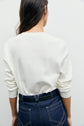 A person with long brown hair, seen from behind, wears a white tee-shirt Bara en coton épais à manches longues, tucked into high-waisted blue jeans with visible stitching and a black belt.