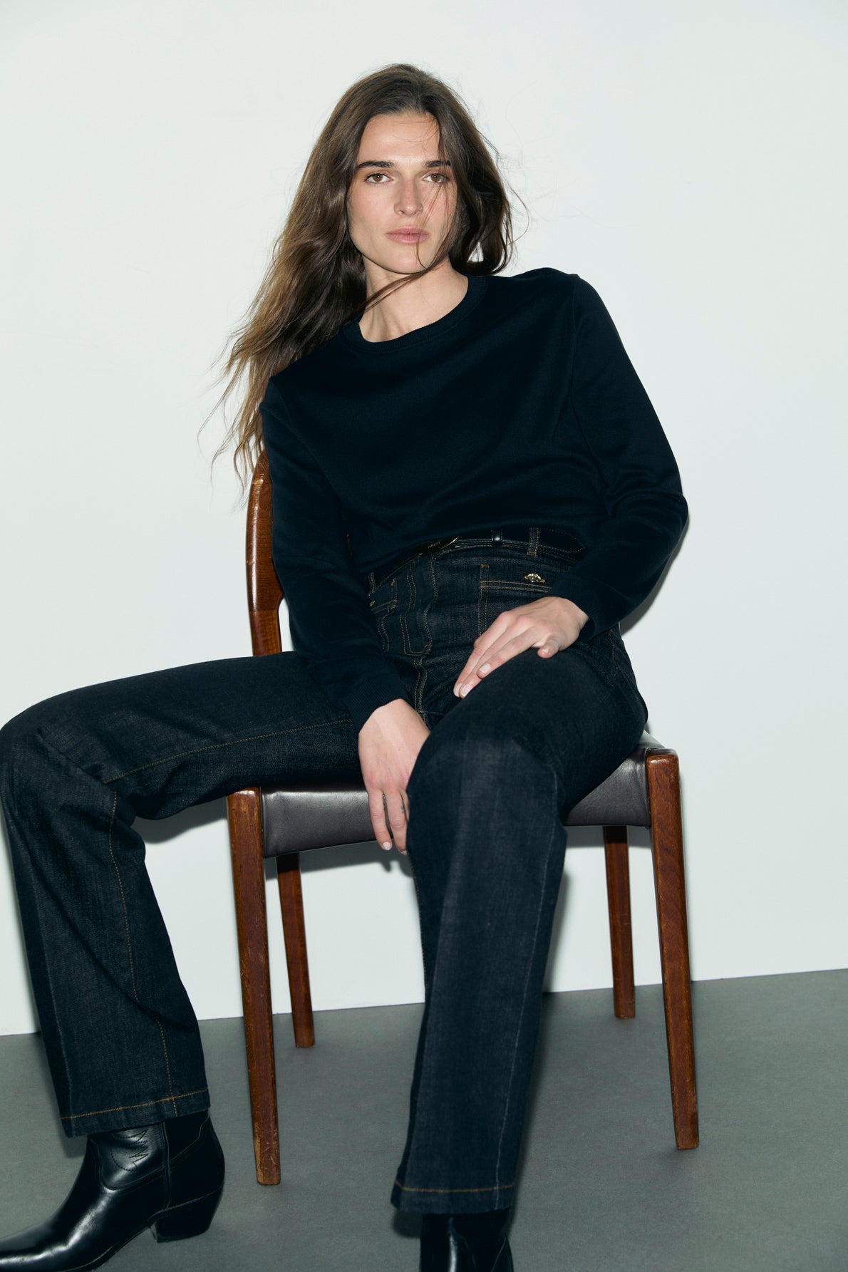 A person with long brown hair sits on a wooden chair, wearing an Esprit sportswear black long-sleeve shirt, dark jeans, and black boots, against a plain white wall and gray floor.