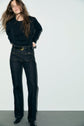 A person with long brown hair wears a black long-sleeve shirt reminiscent of Esprit sportswear, dark high-waisted jeans with front pockets, a black belt, and black shoes, standing against a plain light background.