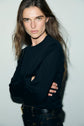 A person with long brown hair wearing a black coton épais sweater and dark jeans stands against a light background, arms crossed and looking directly at the camera with a serious expression, effortlessly embodying an Esprit sportswear vibe.