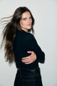 A person with long brown hair and a black tee-shirt Bara in coton épais stands against a white wall, turning slightly to the side with a soft smile and their hair flowing gently.