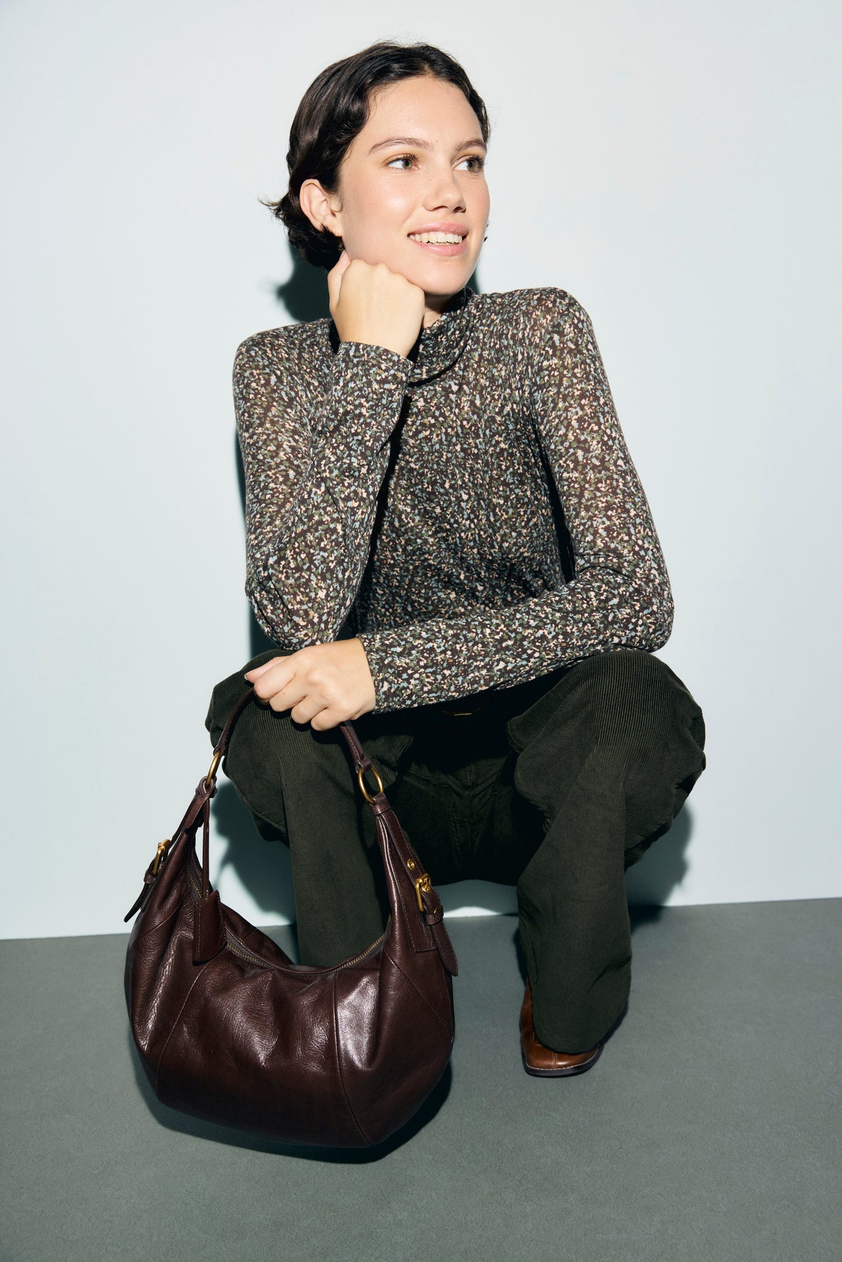 A woman with short brown hair squats, smiling, wearing a t-shirt col roulé à motif imprimé and dark pants. She rests her chin on her hand and has a large brown leather handbag in front of her.