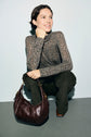 A woman with short brown hair squats, smiling, wearing a t-shirt col roulé à motif imprimé and dark pants. She rests her chin on her hand and has a large brown leather handbag in front of her.