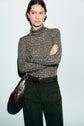 A person wearing a speckled t-shirt col roulé sweater and dark pants stands with arms crossed, carrying a brown shoulder bag, against a light background.