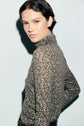 Person with short dark hair wearing a t-shirt col roulé à motif imprimé stands against a light background, looking at the camera with a neutral expression.