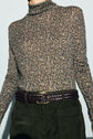 Person wearing a long-sleeve, multicolored t-shirt col roulé with a motif imprimé, dark green corduroy pants, and a dark braided leather belt with a gold buckle. The photo is cropped above the nose.