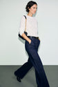 A woman wearing a white lace top with dentelle motifs floraux, dark wide-leg jeans, and black shoes walks while carrying a black purse with a gold chain strap over her shoulder, against a plain light background.