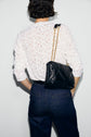 A person wearing a tee-shirt blanc with dentelle motifs floraux and dark jeans stands with their back to the camera, carrying a black quilted purse with a gold chain strap over their shoulder.