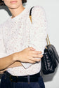 A person in a white, textured, long-sleeve blouse featuring délicats dentelle motifs floraux and dark jeans holds their arm, wearing a black leather quilted bag with a gold chain strap over their shoulder. Their face is partially out of frame.