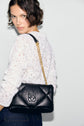 Woman with short brown hair wearing a tee-shirt blanc in matière stretch adorned with dentelle motifs floraux and dark jeans, holding a black quilted shoulder bag with a gold chain strap and a raised logo, standing against a plain light background.