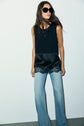 Woman in large black sunglasses, a sleeveless satin top dentelle with lace trim, and light blue flared jeans stands against a plain white wall.