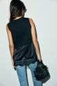 A woman with long dark hair, wearing a sleeveless black top satin with lace trim and light blue jeans, stands facing away, holding a black handbag in her left hand.