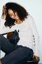 A woman with curly hair, wearing a white tee-shirt coton mailles côtelé and dark jeans, sits smiling with one hand in her hair. A small black quilted purse hangs from her neck.