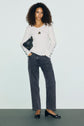 Woman with curly hair wearing a white ribbed sweater in mailles côtelé, loose dark jeans, and black flats stands against a plain light background, holding a small black bag and wearing a necklace.