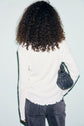 Person with curly hair, wearing a white tee-shirt coton à mailles côtelé and gray jeans, stands facing away from the camera, holding a small black purse over their shoulder against a light background.