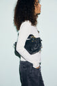 A person with curly hair, wearing a white long-sleeve top featuring delicate dentelle col details and black jeans, stands in profile holding a black woven handbag under their arm against a light blue background.