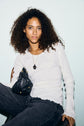 A person with curly hair wearing a white mailles côtelé sweater and dark jeans holds a small black quilted bag. They have a necklace with a black pendant and are sitting against a light background.