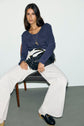 Woman with long dark hair sits on a chair, wearing a navy tee-shirt manches longues in soft coton modal, white wide-leg pants, black loafers, and a gold necklace, holding a black handbag across her lap against a plain background.