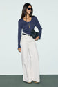 Woman in sunglasses wearing a navy tee-shirt manches longues in soft coton modal, white wide-leg pants, and beige shoes, holding a black textured handbag and wearing a gold necklace, standing against a pale background.