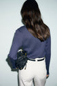 A woman with long brown hair, wearing a ribbed navy tee-shirt coton modal and white pants, stands facing away, holding a black woven handbag under her arm against a light background.