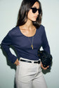 Woman with long brown hair wearing black sunglasses, a dark ribbed tee-shirt manches longues top, white pants, and a black belt, holds a black quilted handbag and wears a gold necklace with a lock pendant.